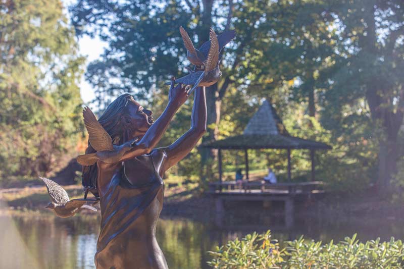 Statue of woman reaching out with birds flying around her in front of the lake.