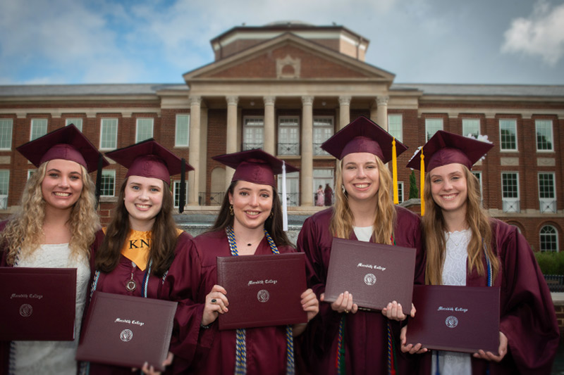 Five graduates smiling in front of Johnson Hall.