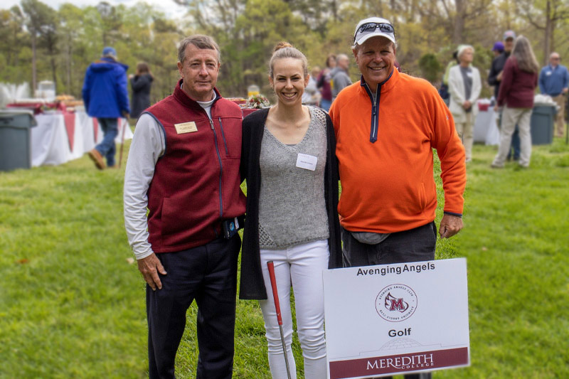 Three people smile behind the Meredith Golf sign.