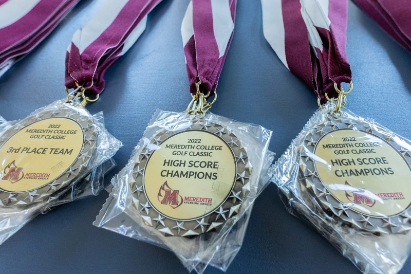 A close up on the medals for the golf tournament that say "2022 Meredith College Golf Classic: High Score Champion" or "3rd Place Team".