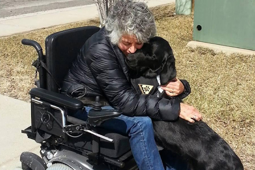 A woman in an electric wheelchair hugging her service dog, a black lab.