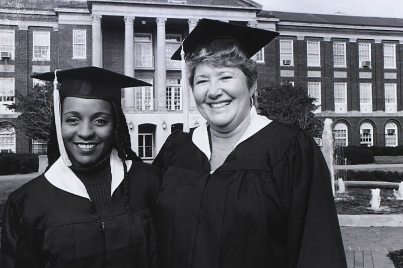 Two wings students in black and white outside Johnson Hall in their graduation cap and gown.