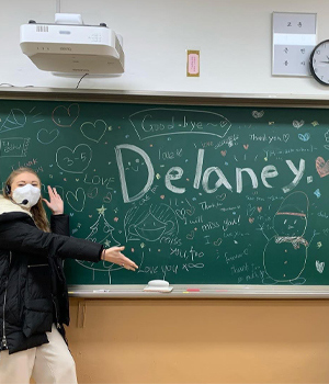A student in a mask shows off her chalkboard with her name, Delaney, written on it and lots of goodbye pictures and messages.