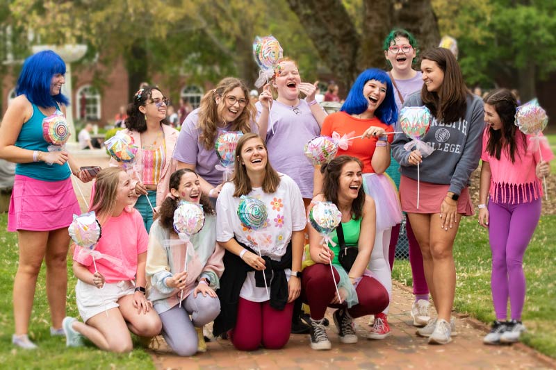 Students at C S A Day laughing wearing bright colors and wigs and holding balloon lollipops.