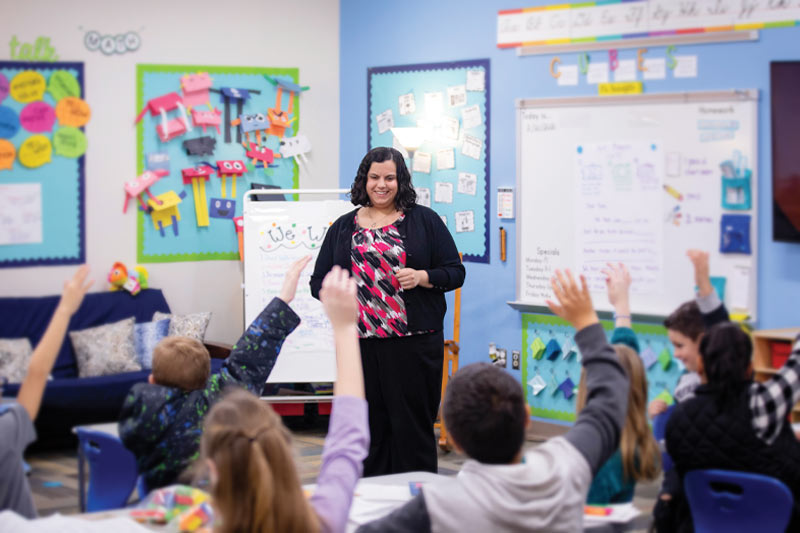 A student smiles as she teaches in a classroom and all her students have their hands raised.
