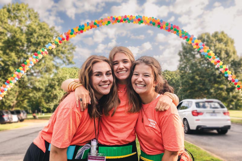 Three upperclassmen smiling as they welcome students in on the main drive; the balloon arch is behind them and framing them from above.