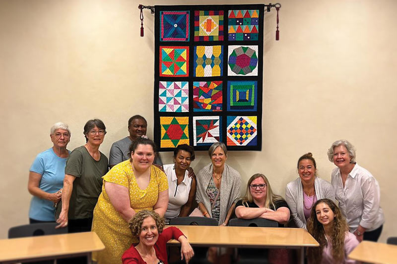 A group of students and other people involved in the quilt project stand in front of a quilt.